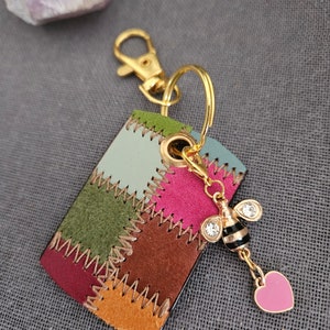 Puede incluir: Un llavero de cuero patchwork colorido con un anillo de metal dorado y un dije con una abeja con pedrería y un corazón rosa.