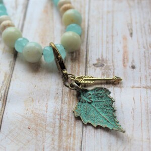 Op de afbeelding: Een turquoise en witte kralenketting met een gouden bladeren hanger en een pijl hanger.