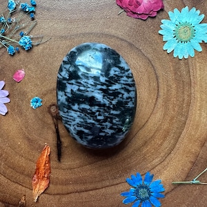 May include: An oval-shaped, polished stone with a black and white pattern, surrounded by dried flowers in various colors. The stone is the focal point, set against a wooden surface.