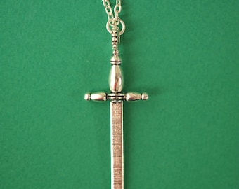 Pode incluir: Um colar com um pingente de espada de prata em uma corrente de prata. A espada tem uma ponta pontiaguda e uma guarda cruzada. O colar está sobre um fundo verde.
