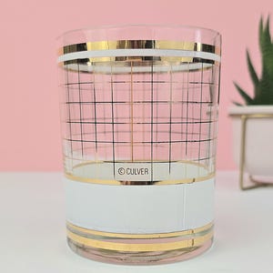 May include: A clear glass tumbler with gold and white bands, and a black grid pattern. The word "CULVER" is printed on a white band. The glass is on a white surface with a pink background.