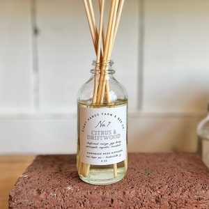 May include: A clear glass bottle with a white label that reads "Eight Hands Farm & Bee Co. No. 7 Citrus & Driftwood Driftwood mango, fig, berry, pineapple, aquatic, mandarin Handmade Reed Diffuser New Hope, PA - Lambertville, NJ 4 oz". The bottle is filled with a clear liquid and has wooden sticks sticking out of the top.