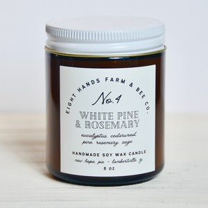 May include: A brown glass jar candle with a white lid. The label reads "Eight Hands Farm & Bee Co. No. 4 White Pine & Rosemary" and lists the scent notes as "eucalyptus, cedarwood, pine, rosemary, sage". The label also states "Handmade Soy Wax Candle" and "New Hope, PA - Lambertville, NJ". The candle is 8 oz.