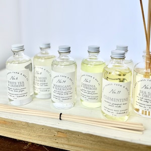 May include: Six glass bottles of essential oil diffuser blends with labels that include the product name and number. The bottles are arranged in a row on a white wooden surface with wooden diffuser sticks.