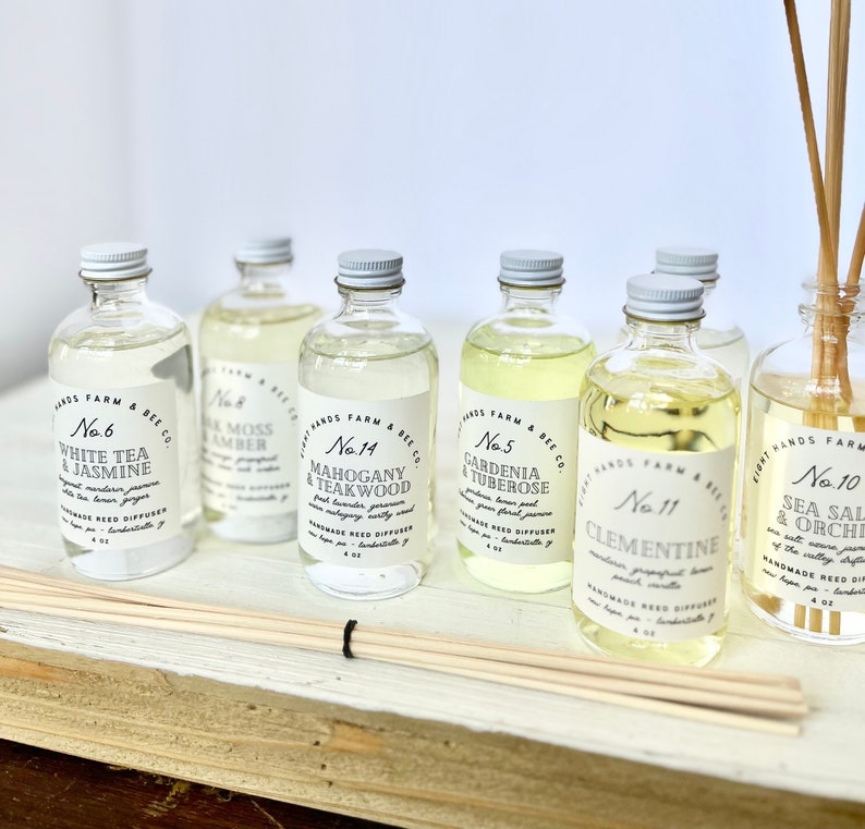 May include: Six glass bottles of essential oil diffuser blends with labels that include the product name and number. The bottles are arranged in a row on a white wooden surface with wooden diffuser sticks.