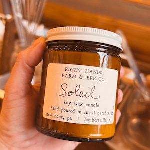 May include: A glass jar candle with a white lid and a label that reads "EIGHT HANDS FARM & BEE CO. Soleil soy wax candle." The candle has a warm, amber color and is hand-poured in small batches.