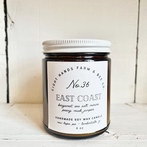 May include: A brown glass jar candle with a white lid and label. The label reads "EIGHT HANDS FARM & BEE CO. No. 36 EAST COAST bergamot, sea salt, marine, peony, musk, juniper HANDMADE SOY WAX CANDLE new hope, pa - lambertielle 8 OZ".