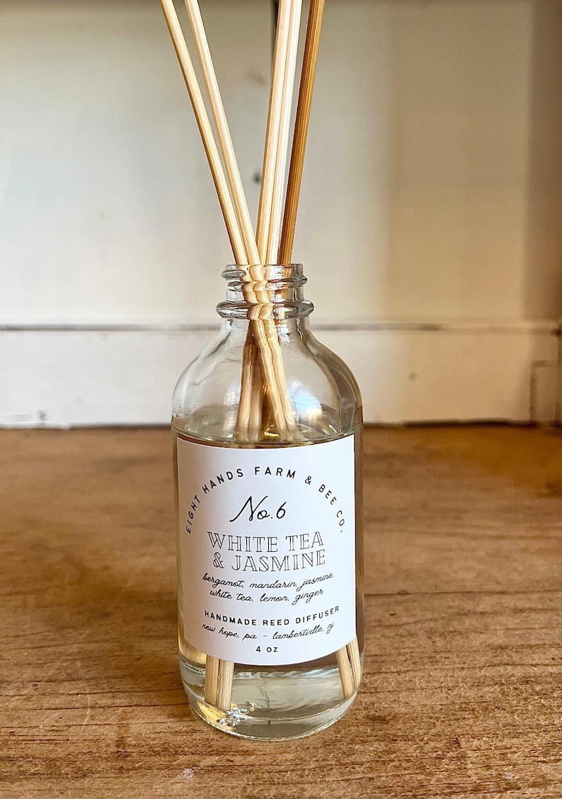 May include: A clear glass bottle with a white label that reads "Light Hands Farm & Bee Co. No. 6 White Tea & Jasmine bergamot, mandarin, jasmine, white tea, lemon, ginger Handmade Reed Diffuser new hope, pa - lambertiville, nj 4 oz". The bottle is filled with a clear liquid and has wooden sticks sticking out of the top.