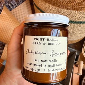 May include: An amber glass jar soy wax candle with a white screw-top lid. The label reads EIGHT HANDS FARM & BEE CO. Autumn Leaves, soy wax candle, hand poured in small batches, new hope, pa | lambertville, nj, and 8 oz.