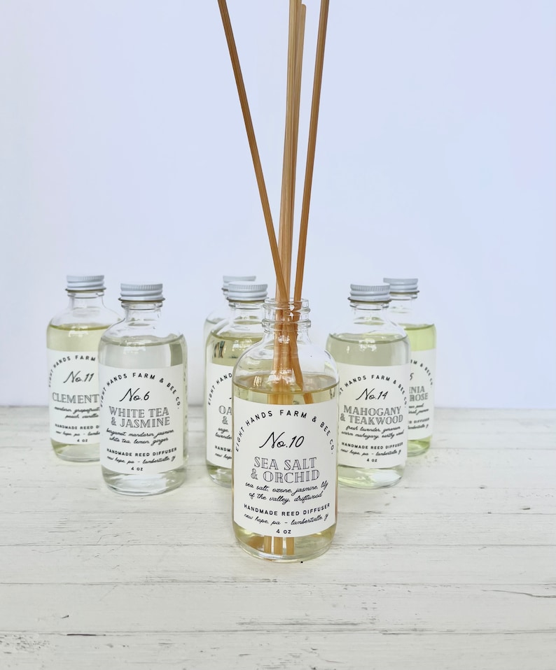 May include: A collection of eight glass bottles with white labels and black text. The bottles contain reed diffusers with various scents, including Sea Salt & Orchid, White Tea & Jasmine, and Mahogany & Teakwood. Each bottle is 4 oz and features a handwritten label with the scent name and number.