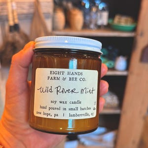 May include: A brown glass jar candle with a white lid. The label reads "Wild River Mint" and "soy wax candle." The candle is hand-poured in small batches by Eight Hands Farm & Bee Co. in New Hope, PA and Lambertville, NJ. The jar is held in a person's hand.