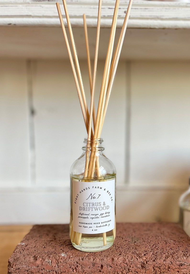 May include: A clear glass bottle with a white label that reads "Eight Hands Farm & Bee Co. No. 7 Citrus & Driftwood Driftwood mango, fig, berry, pineapple, aquatic, mandarin Handmade Reed Diffuser New Hope, PA - Lambertville, NJ 4 oz". The bottle is filled with a clear liquid and has wooden sticks sticking out of the top.