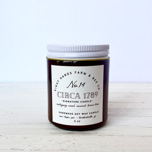 May include: A brown glass candle jar with a white lid. The label reads "Eight Hands Farm & Bee Co. No. 14 Circa 1789 - Signature Candle - mahogany wood, coconut, lemon-lime Handmade Soy Wax Candle new hope, pa - lambertielle, ga 8 oz"