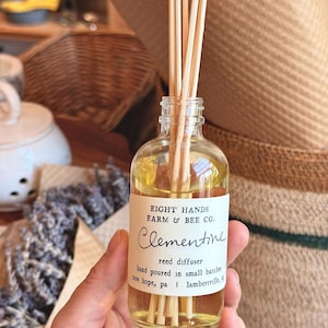 May include: A clear glass reed diffuser bottle filled with yellow liquid and several wooden reeds. The white label reads Eight Hands Farm & Bee Co. Clementine reed diffuser, hand poured in small batches, New Hope, PA | Lambertville, NJ.
