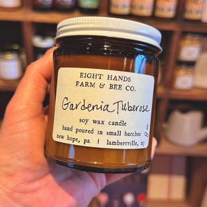 May include: A brown glass jar with a white lid, labeled "Gardenia Tuberose" soy wax candle from Eight Hands Farm & Bee Co. The label indicates the candle is hand-poured in small batches in New Hope, PA and Lambertville, NJ.