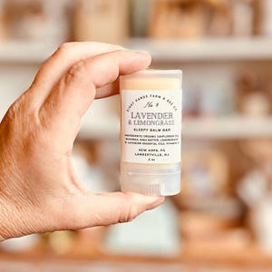 May include: A white tube with a clear top containing a lavender and lemongrass sleepy balm bar. The tube is labeled "Eight Hands Farm & Bee Co. No. 9 Lavender & Lemongrass Sleepy Balm Bar" and lists the ingredients as "Organic Sunflower Oil, Beeswax, Shea Butter, Lemongrass & Lavender Essential Oils, Vitamin E". The tube also lists the location as "New Hope, PA Lambertville, NJ" and the size as "0.5 oz."