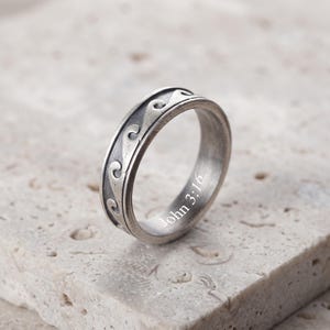 Op de afbeelding: Een zilverkleurige metalen ring met een verhoogd golfpatroon rond de buitenste band. Het binnenoppervlak van de ring is gegraveerd met de tekst John 3:16 in een strak, wit lettertype. De ring heeft een geborstelde, geoxideerde afwerking.