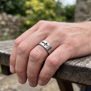 Peut inclure: Une bague anneau en métal argenté présentant un motif de croix découpé. La bague possède une finition mate lisse et un profil plat, entourant le doigt avec une esthétique simple et épurée qui met en valeur l'espace négatif de la croix.