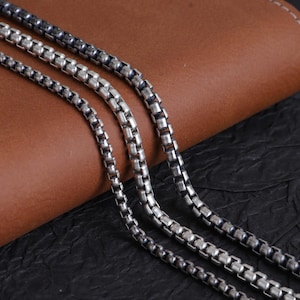 Puede incluir: Primer plano de tres collares de cadena de caja plateados. Las cadenas se muestran sobre una superficie de cuero marrón con un fondo negro. Las cadenas tienen un diseño de eslabones cuadrados, con una cadena con un acabado más oscuro.