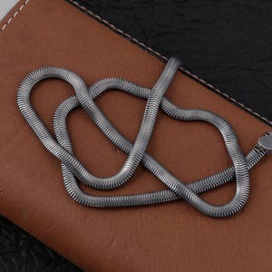 May include: A silver snake chain necklace with a flat, flexible design. The necklace is displayed on a brown leather surface with a dark, textured background. The chain's links create a textured, fluid appearance.