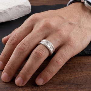 May include: A wide silver band ring featuring a repeating pattern of engraved geometric symbols around the entire exterior surface. The metal has a matte finish and a textured appearance, with a consistent width and a slightly rounded profile.