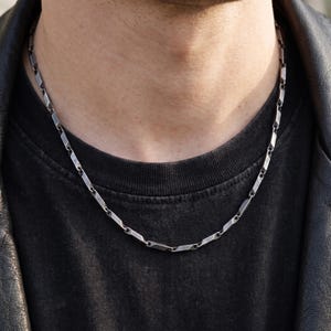 May include: A silver-toned metal chain necklace featuring a series of elongated, rectangular links with beveled edges. The chain has a polished finish and rests against a black t-shirt and a black leather jacket, showcasing a structured design.