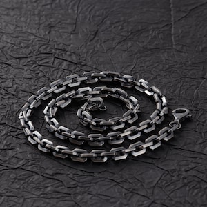 Puede incluir: Un collar de cadena plateada con un diseño de eslabones rectangulares. El collar se muestra sobre una superficie oscura y texturizada. La cadena está enrollada, mostrando el cierre y el intrincado patrón de eslabones.