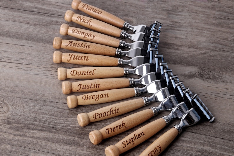 May include: A collection of personalized razors with wooden handles and engraved names. The razors have silver-colored heads and are arranged on a wooden surface. Each handle is engraved with a different name.