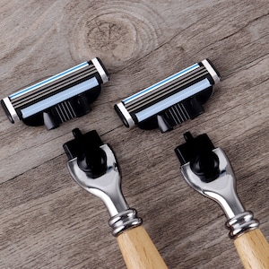 May include: Close-up of two razors with wooden handles and four replacement razor heads. The razor heads have multiple blades with blue and silver accents. The razors and heads are on a wooden surface.