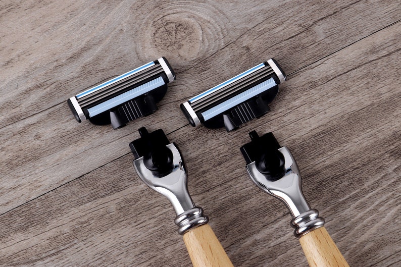 May include: Close-up of two razors with wooden handles and four replacement razor heads. The razor heads have multiple blades with blue and silver accents. The razors and heads are on a wooden surface.