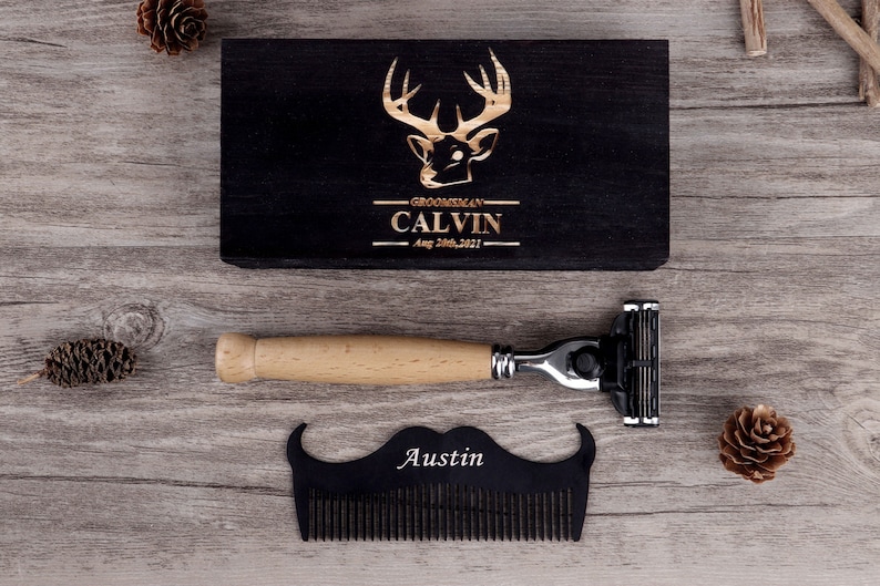 May include: A black wooden box with a deer head design and the text "Groomsman CALVIN Aug 28th, 2021". A razor with a wooden handle and a black mustache-shaped comb with the name "Austin" are also present.