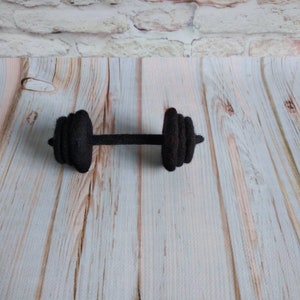 May include: A black felt dumbbell with a textured surface. The dumbbell is lying on a light wood surface.
