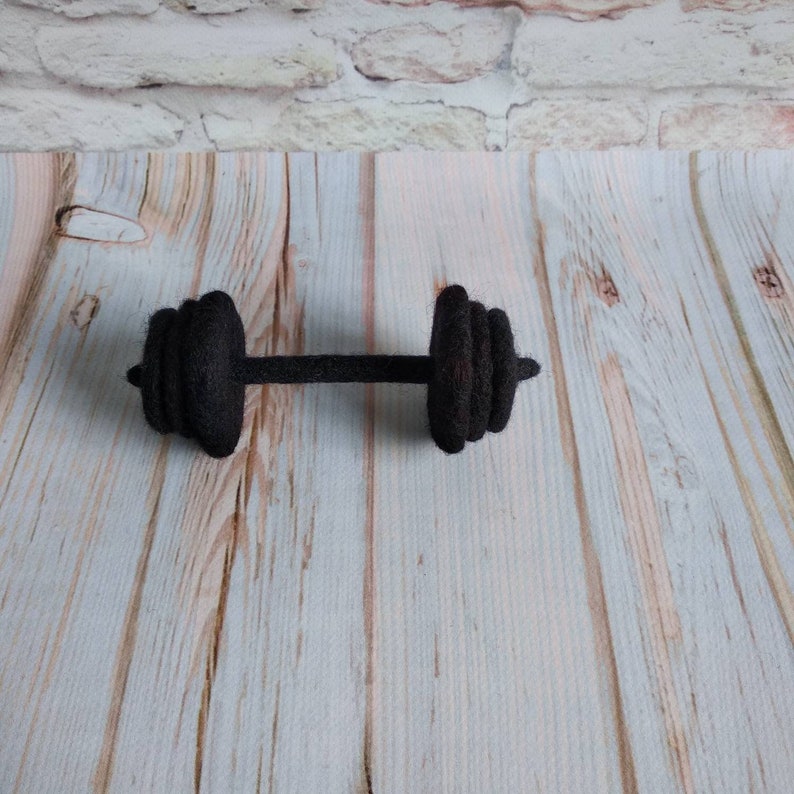 May include: A black felt dumbbell with a textured surface. The dumbbell is lying on a light wood surface.