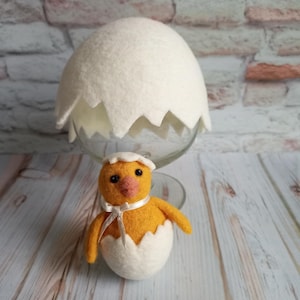 May include: A yellow felt chick with a white felt eggshell, peeking out of the top of the eggshell. The chick has black eyes and a white ribbon around its neck. The eggshell is sitting on a clear glass pedestal.