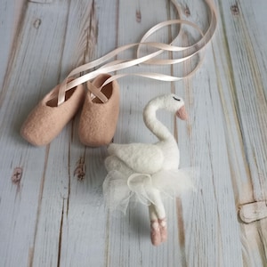 Puede incluir: Una figura de cisne de fieltro con tutú blanco y zapatillas de ballet. El cisne tiene el cuello curvado y los ojos cerrados. Las zapatillas de ballet son de color rosa claro con cintas a juego.