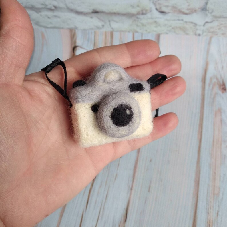 May include: A small gray and white felt camera with a black lens. The camera has a black strap attached to it.