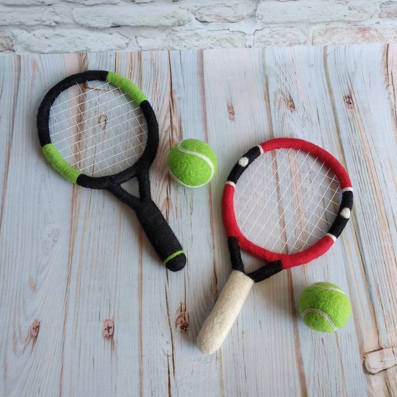 K&ouml;nnte beinhalten: Zwei Filz-Tennisschl&auml;ger und zwei Filz-Tennisb&auml;lle auf einer Holzoberfl&auml;che. Ein Schl&auml;ger ist schwarz und gr&uuml;n, der andere ist rot und schwarz. Die B&auml;lle sind gr&uuml;n.