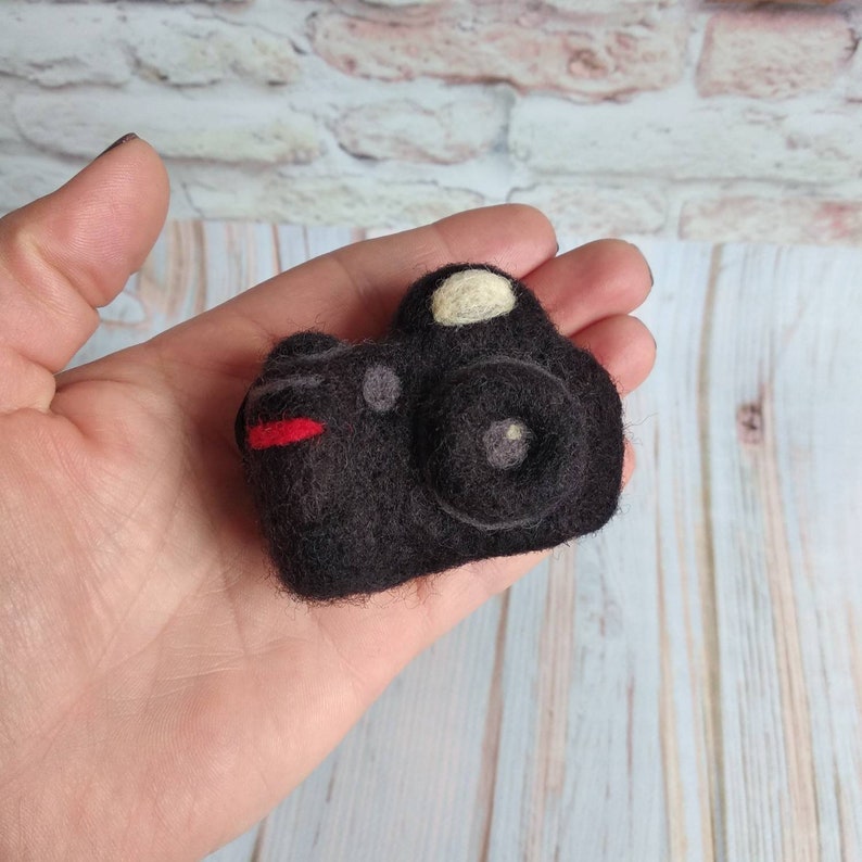 May include: A black felt camera with a red stripe and a white lens. The camera is handmade and has a vintage look.