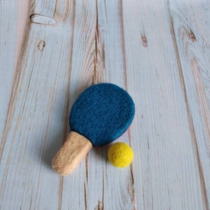 K&ouml;nnte beinhalten: Ein blaues und braunes Filz-Tischtennisblatt und ein gelber Filzball auf hellem Holzgrund.