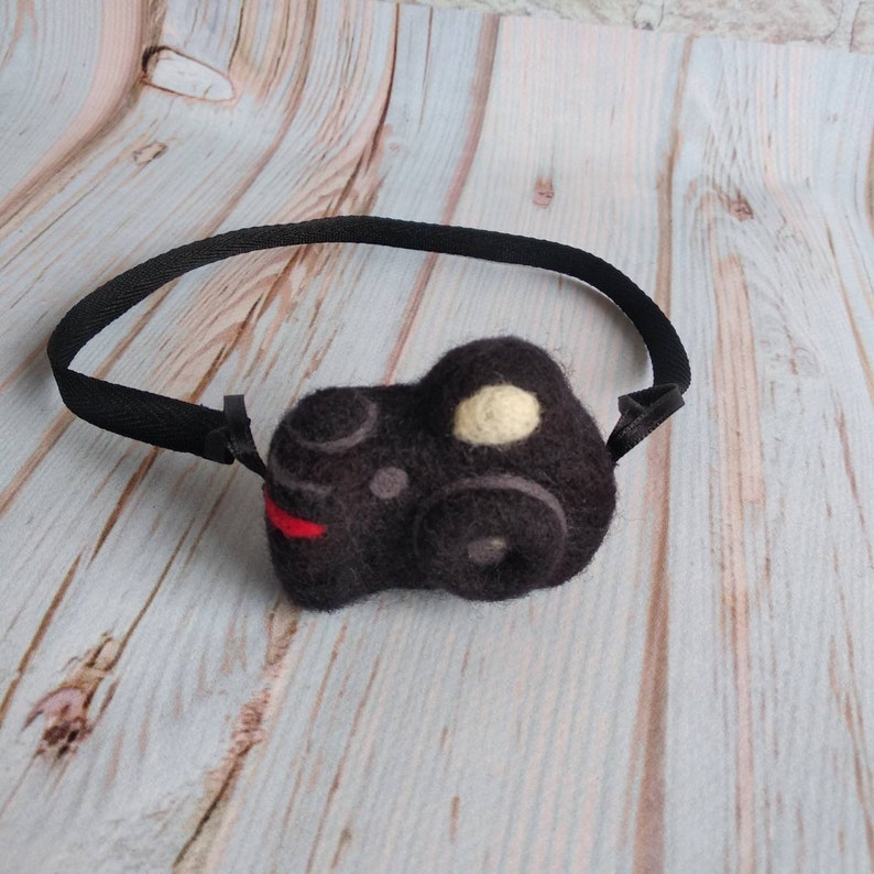 May include: A black felt camera headband with a white circle detail. The headband is made of a soft, black felt material and has a black elastic band.