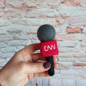 以下が含まれることがあります： 黒いハンドルと赤いボディに白い文字「CNN」が書かれたフェルト製の玩具マイクです。