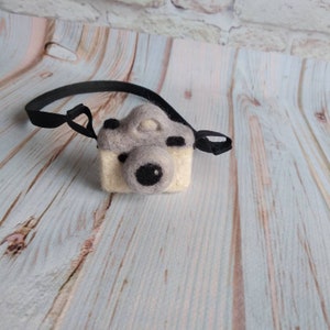May include: A gray and white felt camera with a black strap. The camera has a black felt lens and a white felt body.