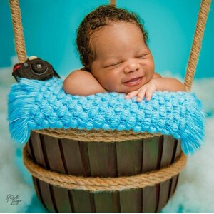 May include: A newborn baby boy is sleeping in a blue and brown basket with a plush toy. The basket is decorated with a rope trim.