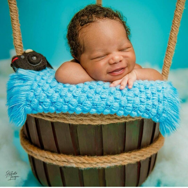 May include: A newborn baby boy is sleeping in a blue and brown basket with a plush toy. The basket is decorated with a rope trim.