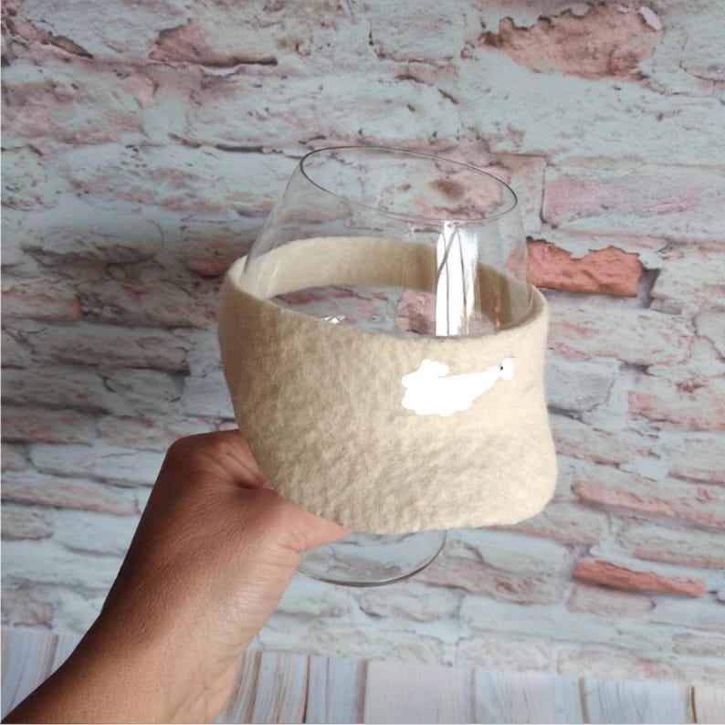 May include: A clear glass vessel wrapped in a textured, beige-colored fabric sleeve. The sleeve features a small, white, irregular-shaped patch on the front surface. The glass has a wide, rounded bowl shape and a short, sturdy base for stability.