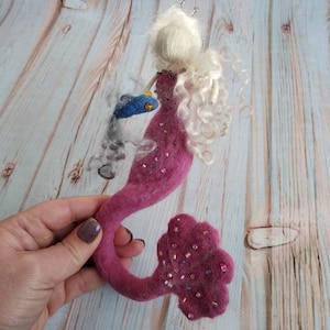 Könnte beinhalten: Eine rosa Filz-Meerjungfrau mit einer weißen Filzkrone, langen weißen Haaren und einem blauen Filzfisch. Die Meerjungfrau ist mit rosa und goldenen Perlen verziert.