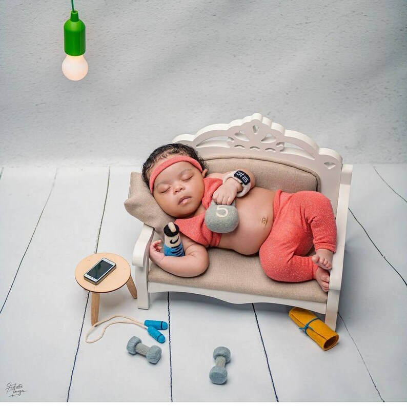 May include: A baby wearing a red outfit and a watch is sleeping on a white couch. The baby is holding a small gray weight with the number 5 on it. There are blue and gray dumbbells and a jump rope on the white wooden floor.