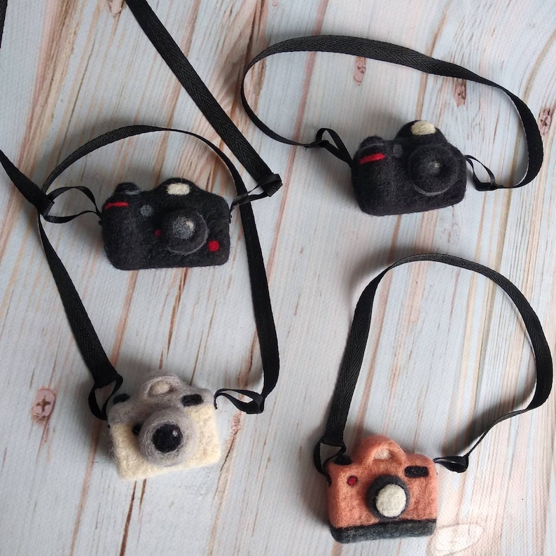 May include: Four felt camera keychains with black straps. The cameras are black, gray, and brown with white details.