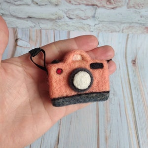 May include: A small, peach-colored felt camera with a black strap and a white lens. The camera is held in a hand.