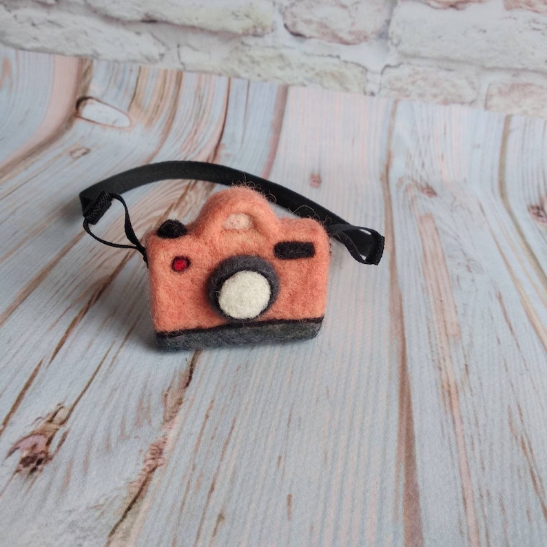 May include: A small, felt camera with a black strap. The camera is peach colored with a black lens and a white circle in the center.